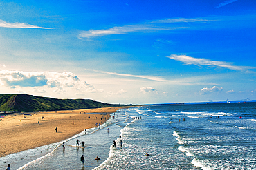 Sunny beach scene with people and rolling waves, blue sky with clouds overhead, and a hilly backdrop in Saltburn-by-the-Sea, England.