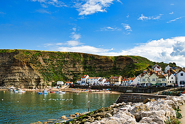 Quaint coastal village with white houses nestled against a cliff, overlooking a serene harbor with boats under a blue sky with clouds in Staithes, England.