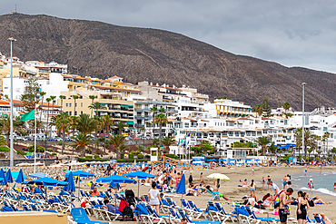 Crowded beach with tourists, sun loungers, and umbrellas against a backdrop of a mountain and coastal town in Los Cristianos, Tenerife, Canary Islands, Spain, Atlantic, Europe