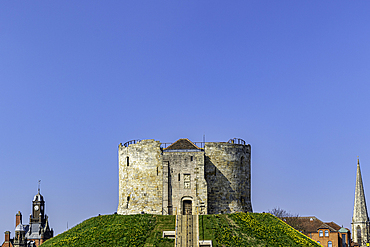 Medieval stone tower on a grassy hill with clear blue sky, flanked by church spires in the background, Clifford's Tower, York, North Yorkshire, England, United Kingdom, Europe