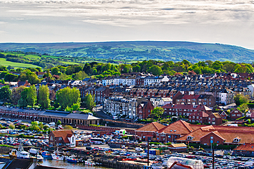 Aerial view of a coastal town with a harbor filled with boats. The town features a mix of residential buildings and greenery, with rolling hills in the background under a partly cloudy sky.