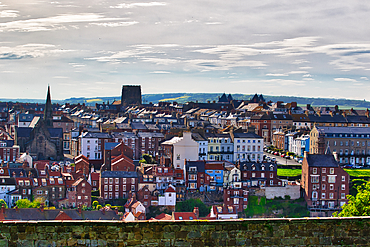 A panoramic view of a coastal town with a mix of historic and modern buildings, under a cloudy sky.