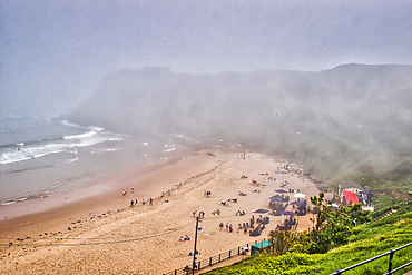 A foggy beach scene with people relaxing on the sand and a few tents set up. The ocean waves are gently crashing onto the shore, and the background is obscured by dense fog.