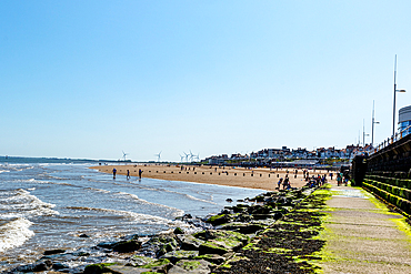 Sunny beach side with people walking along the pier, clear blue sky, and gentle waves lapping against the shore in Bridlington, Yorkshire, England, United Kingdom, Europe