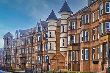 A row of modern brick apartment buildings with unique cylindrical towers and pointed roofs. The buildings have multiple windows and black metal fences in front.