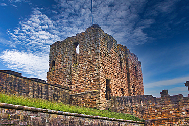 An ancient stone castle ruin with a partially collapsed tower, set against a blue sky with scattered clouds. The structure is surrounded by a stone wall and grassy area.