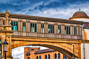 A pedestrian bridge with glass windows connecting two buildings under a cloudy sky. The bridge has an architectural design with statues and ornate details.