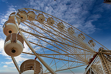 A close-up view of a large Ferris wheel against a blue sky with clouds. The structure features white metal beams and circular cabins, creating a striking visual against the backdrop.