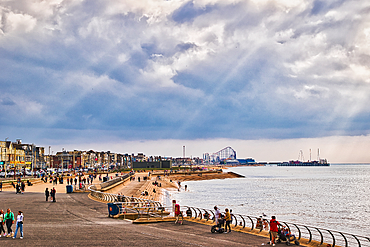 A scenic view of a beach promenade with people walking along the shore. The sky is partly cloudy with rays of sunlight breaking through. In the background, a pier and amusement rides are visible, adding to the coastal atmosphere.