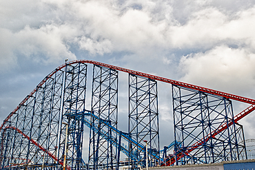 A towering roller coaster structure with a red and blue track against a cloudy sky. The intricate metal framework supports the thrilling ride, showcasing its height and design.