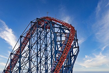 A towering roller coaster track ascending into a clear blue sky with wispy clouds, showcasing the intricate steel structure and vibrant red track.