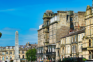 A scenic view of historic buildings in a town square, featuring ornate architecture and a tall monument under a clear blue sky.