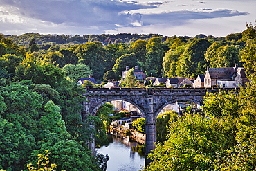 A scenic view of a stone bridge arching over a river, surrounded by lush greenery and quaint houses. The sky is partly cloudy, adding depth to the landscape.