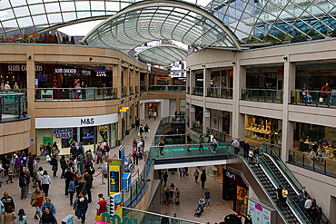 A bustling shopping mall with multiple levels, featuring various retail stores and shoppers. The architecture includes a glass roof and open walkways, creating a vibrant atmosphere.