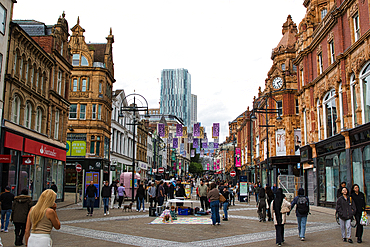 A bustling urban street scene featuring historic architecture, shops, and pedestrians. The street is lively with people walking, and colorful banners hang overhead. Modern buildings are visible in the background, contrasting with the ornate facades of the