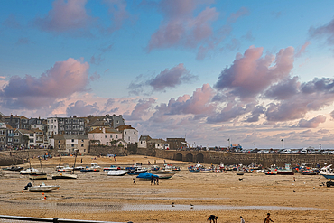 A picturesque coastal scene featuring a sandy beach at low tide, dotted with various boats. In the background, charming houses and a stone arch bridge are visible under a colorful sky with fluffy clouds. People stroll along the beach, enjoying the serene