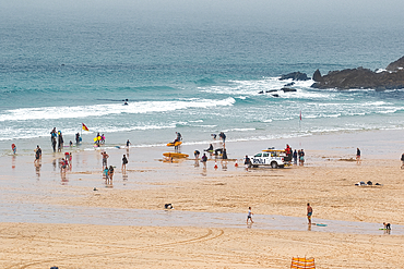 A busy beach scene with people enjoying various activities. Lifeguards are present, and some individuals are surfing in the water. The beach is sandy and wet, with a cloudy sky overhead.