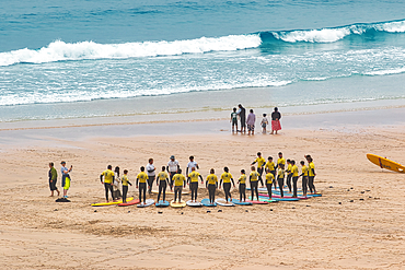 A group of surfers in yellow shirts gathered on the beach, preparing for a surfing lesson. The ocean waves crash in the background, and some people walk along the shore.