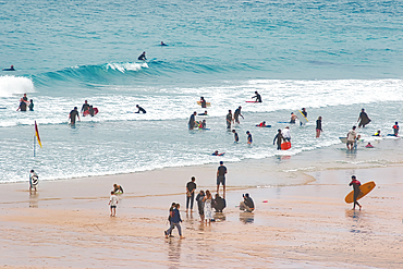 A busy beach scene with people enjoying various activities. Some are surfing, while others are playing in the water or walking along the shore. The waves are gentle, and the atmosphere is lively and cheerful.