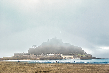 A misty view of a coastal landscape featuring a castle on a hill, surrounded by fog. The foreground shows a beach with people walking, while the background reveals the castle partially obscured by clouds.