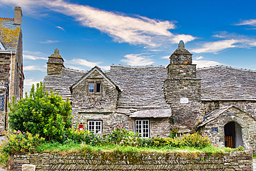 A charming stone cottage with a slate roof, surrounded by lush greenery and a well-maintained garden. The sky is bright with scattered clouds, adding to the picturesque scene.