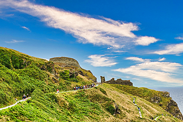 A scenic view of a hillside with lush greenery and a winding path. People are walking along the path near ancient ruins, under a bright blue sky with scattered clouds.