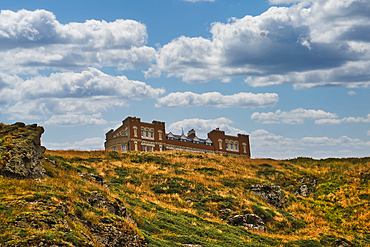 A historic building perched on a grassy hill under a cloudy sky. The structure features ornate architecture with large windows, surrounded by a natural landscape of rolling hills and rocky outcrops.