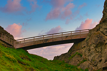 A wooden bridge arches over a rocky landscape, surrounded by lush green grass and cliffs under a colorful sky with soft clouds.