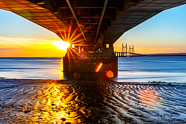 Sunset view under a pier with sunburst effect, reflecting on wet sand and calm water, Prince of Wales Bridge, the Second Severn Crossing, M4 motorway over the River Severn, between England and Wales, United Kingdom, Europe