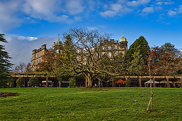 A large stone building with cupolas is partially obscured by a leafless tree in the foreground. A covered walkway with market stalls is visible below the building, leading to a grassy area. The sky is mostly blue with some clouds.