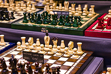 Multiple handmade chess sets displayed on a dark surface. Sets feature various wood types and piece colors, including green, white, and red. Price tags are visible.