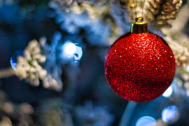 A close-up shot of a single red glitter Christmas ornament hanging on a blurred, snow-covered artificial Christmas tree with out-of-focus blue and white lights.