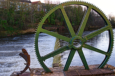 Large green metal gear wheel overlooking a river with a small waterfall and sculpture. Buildings and trees are visible in the background in Wetherby, UK.