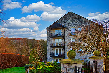 Three-story stone apartment building with balconies, situated in a landscaped garden setting under a partly cloudy blue sky. Stone pillars and a black metal fence are in the foreground in Wetherby, UK.