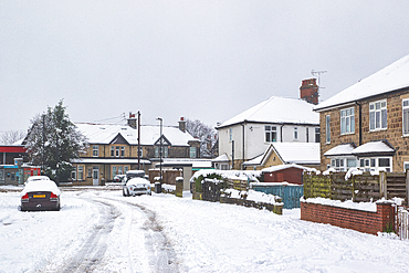 Snow-covered residential street scene with parked cars in front of two-story houses. The houses are various colors and styles, with snow on roofs and fences in Harrogate, UK.