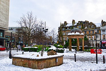 Snow-covered town square with a stone gazebo, buildings, and landscaping. Pedestrians are visible in the background in Harrogate, UK.