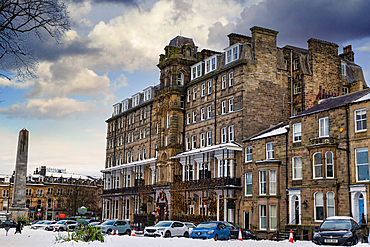 Snow-covered Harrogate town square features a large stone hotel adorned with Christmas lights, adjacent buildings, parked cars, and a tall obelisk in Harrogate, UK.