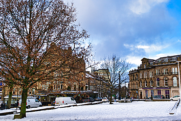 Snow-covered town square with Victorian-era buildings, bare trees, and parked vans. A partly cloudy sky is visible above in Harrogate, UK.