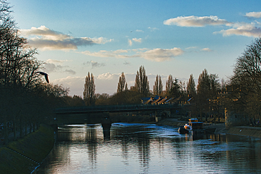 A river scene features a bridge, trees, buildings, a boat, and a bird in flight against a cloudy blue sky in York, UK.