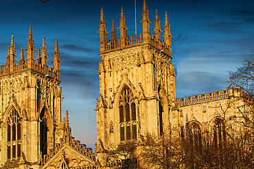 Two ornate, Gothic-style cathedral towers dominate the frame against a blue, slightly cloudy sky. Warm light illuminates the stone structures.