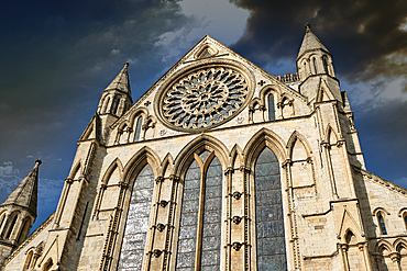 Low-angle shot of a large cathedral facade featuring Gothic architecture, including arched windows, spires, and a rose window, against a cloudy sky.