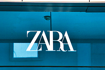 A close-up of the ZARA logo, white, on a teal glass storefront with reflections and a security camera visible in Manchester, UK.