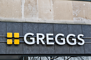 Greggs store sign. White lettering and orange squares on a dark gray tile background, with a lighter stone wall above in Liverpool, UK.