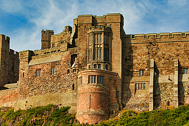 A large, imposing stone castle with multiple towers, walls, and windows, viewed from a low angle. Green vegetation at the base at Bamburgh Castle, UK.