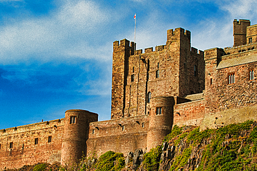 Low-angle view of a large, stone castle with turrets against a blue sky, situated on a rocky outcropping covered in greenery at Bamburgh Castle, UK.