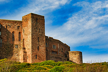 A stone castle complex stands on a grassy hill against a blue sky with scattered white clouds at Bamburgh Castle, UK.