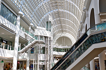 Interior view of a shopping mall with a glass roof, escalators, an elevator, and multi-level storefronts. Mostly white and glass architectural elements at Bentall Centre in Kingston, UK.