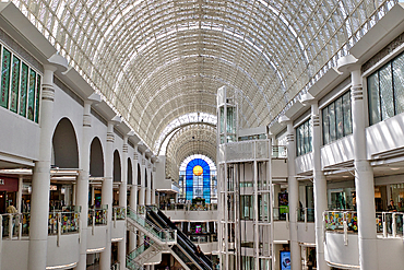 Interior view of a multi-story shopping mall with a vaulted glass ceiling, columns, escalators, and a glass elevator shaft at Bentall Centre in Kingston, UK.