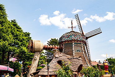 An eye-level shot features a whimsical windmill with faux-wood elements, nestled among green foliage and a partial blue sky at Disneyland Paris.
