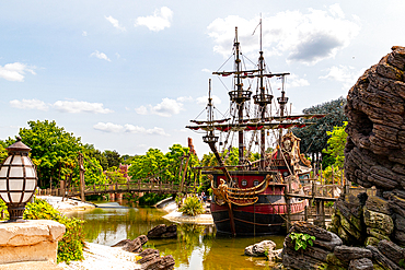 View of a docked pirate ship in a waterway, surrounded by lush greenery, a bridge, a rock formation, and an old-fashioned lantern at Disneyland Paris.